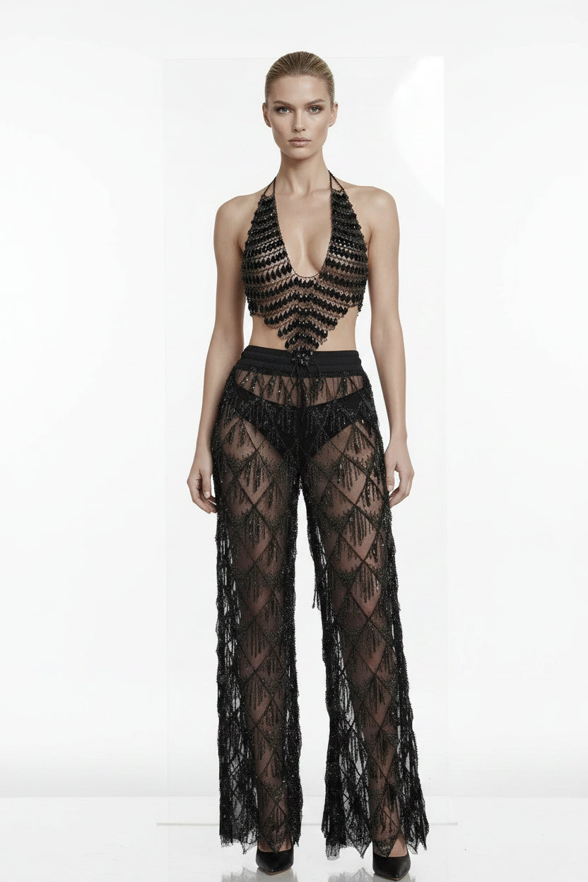 A model wearing a black sheer lace halter crop top and matching wide-leg pants with a geometric diamond lace pattern, set against a plain white background.