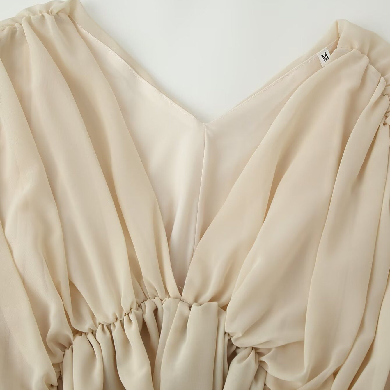 Close-up of a beige or cream-colored draped garment with a V-neckline, gathered elastic details, and soft flowing fabric folds.
