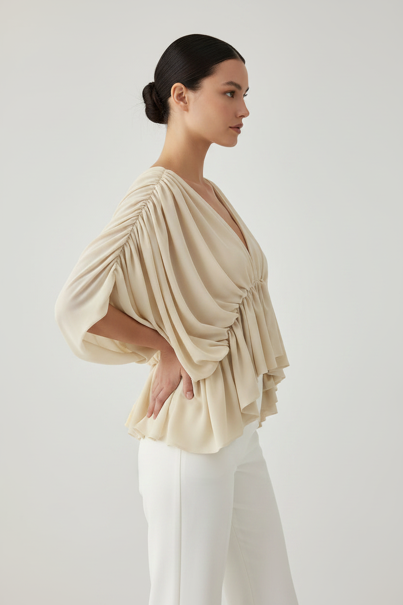 A woman in profile wearing a draped, pleated beige blouse with voluminous elbow-length sleeves and white pants, photographed against a clean light gray studio background.